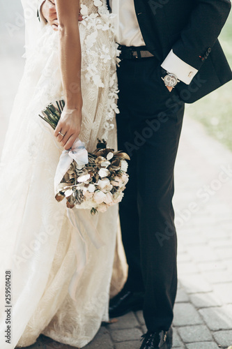 luxury wedding couple hugging and holding golden bouquet of roses with ribbons  in park. stylish groom and gorgeous bride in expensive gown. wedding reception. space for text.
