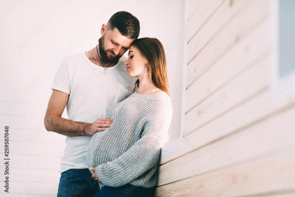 Young pregnant couple near the window of the house, on the background of a brick white wall and a wooden window sill Young pregnant couple near the window of the house, on the background of a brick white wall and a wooden window sill