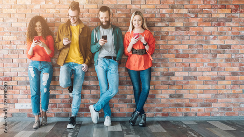 Millennials social media addicted generation. Young people in colorful outfits leaned against brick wall, using smartphones.