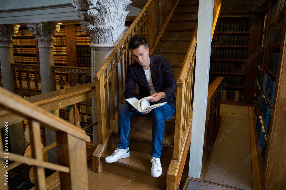 Young handsome man sitting on wooden stairs in traditional library, holding book. Beautiful interior of old library with pillars. Higher education. Young handsome man sitting on wooden stairs in traditional library, holding book. Beautiful interior of old library with pillars. Higher education.