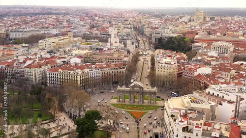 Busy Puerta de Alcalá in Madrid. Cars are driving on the street and pedestrians are entering and leaving the park El Retiro. The scenery is shown in an aerial shot on a semi cloudy day.