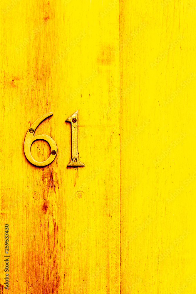 Yellow bright House number 61 with the sixty-one in bronze numbers on a varnished light wooden door with visible grain Yellow bright House number 61 with the sixty-one in bronze numbers on a varnished light wooden door with visible grain