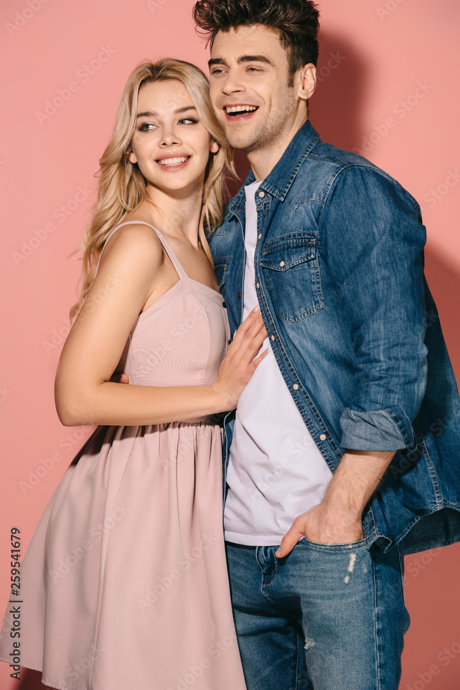 smiling girlfriend in pink dress and handsome boyfriend in denim shirt hugging and looking away smiling girlfriend in pink dress and handsome boyfriend in denim shirt hugging and looking away