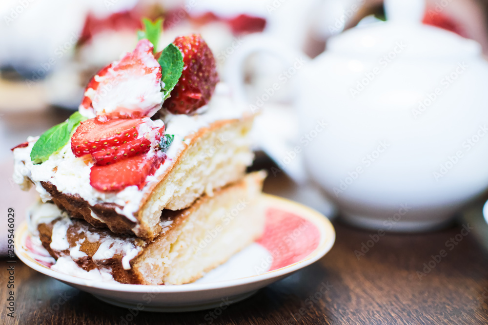 A piece of strawberry cake in cream and mint leaves on a table in a plate, in front of a teapot with tea, in light colors, close-up with a blurred background. A piece of strawberry cake in cream and mint leaves on a table in a plate, in front of a teapot with tea, in light colors, close-up with a blurred background.