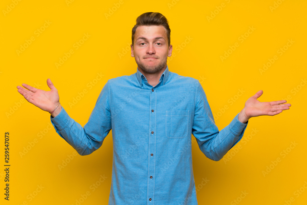 Blonde man over isolated yellow wall having doubts while raising hands Blonde man over isolated yellow wall having doubts while raising hands