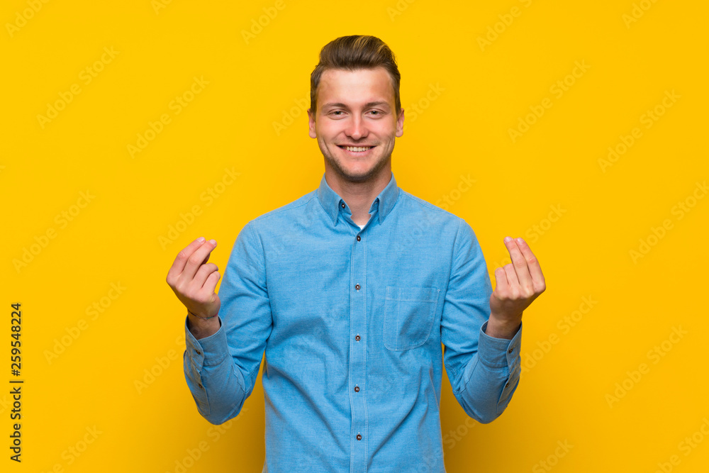 Blonde man over isolated yellow wall making money gesture Blonde man over isolated yellow wall making money gesture