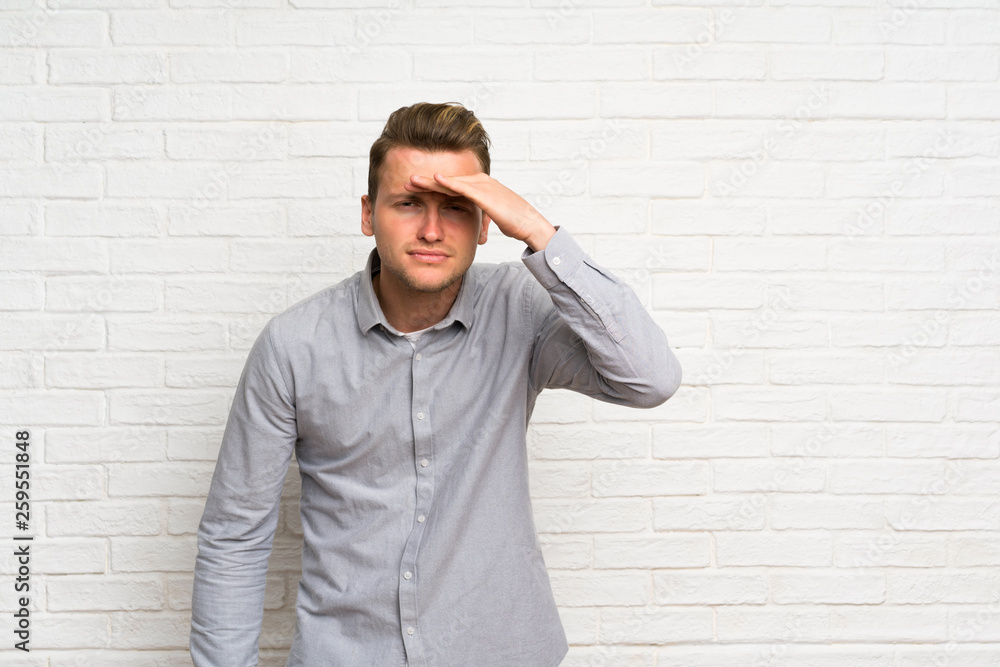 Blonde man over white brick wall looking far away with hand to look something Blonde man over white brick wall looking far away with hand to look something