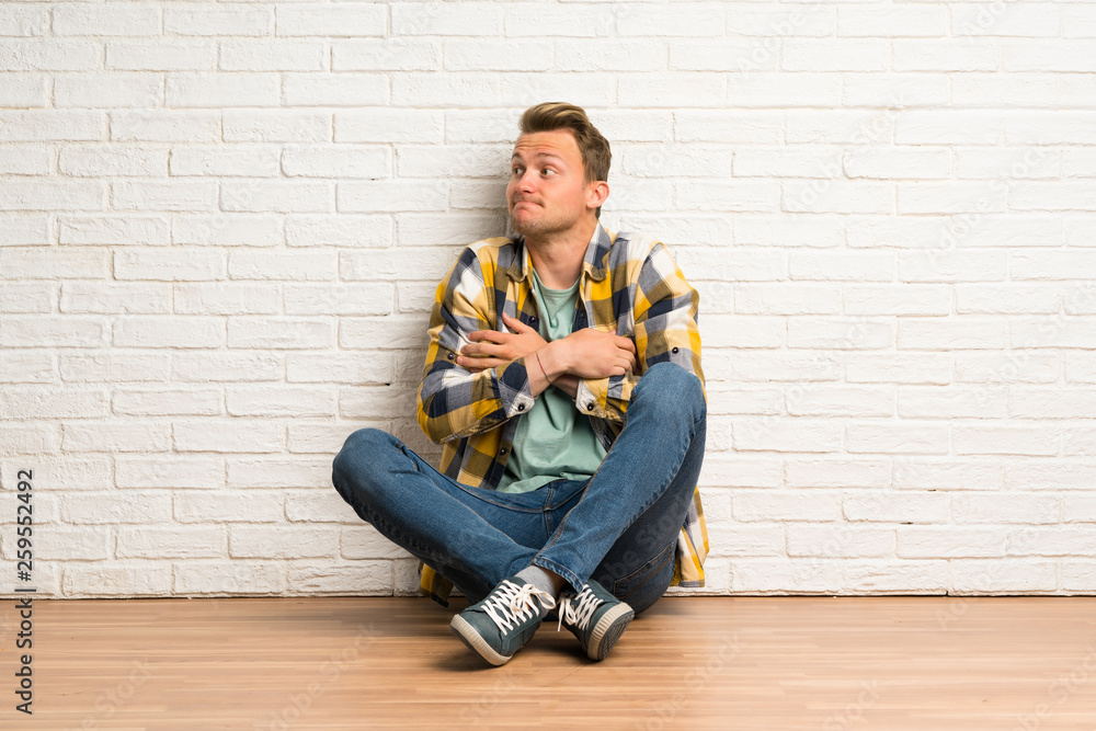 Blonde man sitting on the floor making doubts gesture while lifting the shoulders Blonde man sitting on the floor making doubts gesture while lifting the shoulders