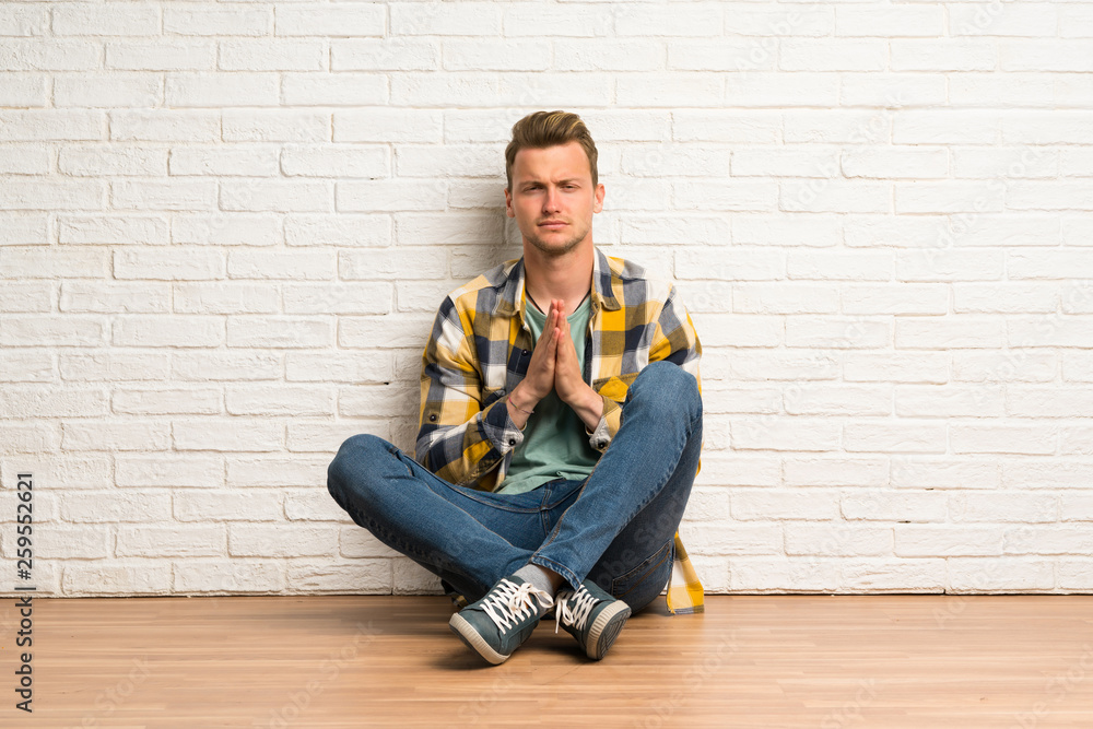 Blonde man sitting on the floor keeps palm together. Person asks for something Blonde man sitting on the floor keeps palm together. Person asks for something