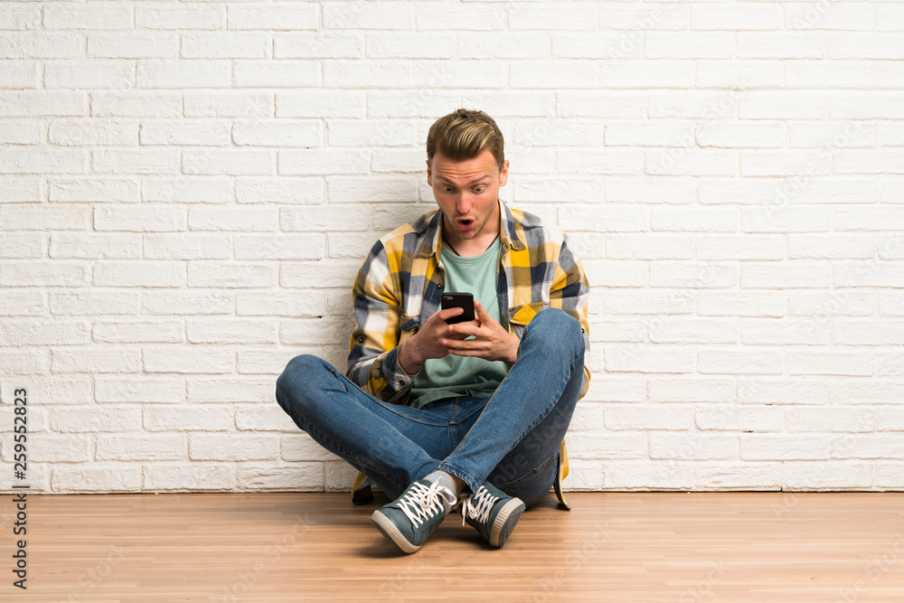Blonde man sitting on the floor surprised and sending a message Blonde man sitting on the floor surprised and sending a message