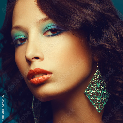 Synth-pop concept. Close up portrait of fashionable model with curly hair and arty make-up posing over blue background. Vintage earrings. Perfect skin. Disco retro style. Studio shot