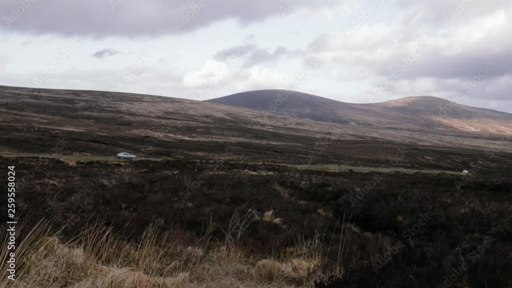 Pan left moody shot as a car driving through the scene, open brown plain and wilderness with mountains and grass that dancing int he wind Pan left moody shot as a car driving through the scene, open brown plain and wilderness with mountains and grass that dancing int he wind