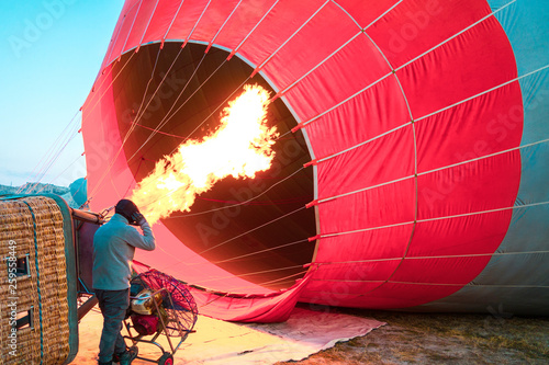 Hot air balloon with flame and basket lying on the ground on the field while filling with air and preparing to take off.