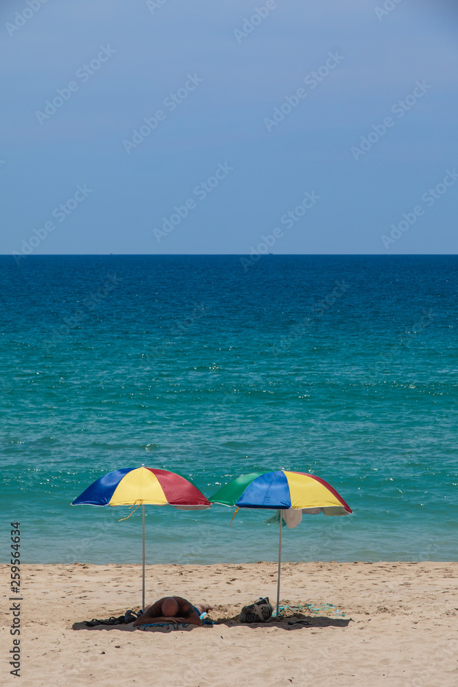 A tourist sleep under two colorful umbrellas on white sand beach.