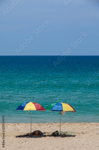 A tourist sleep under two colorful umbrellas on white sand beach.