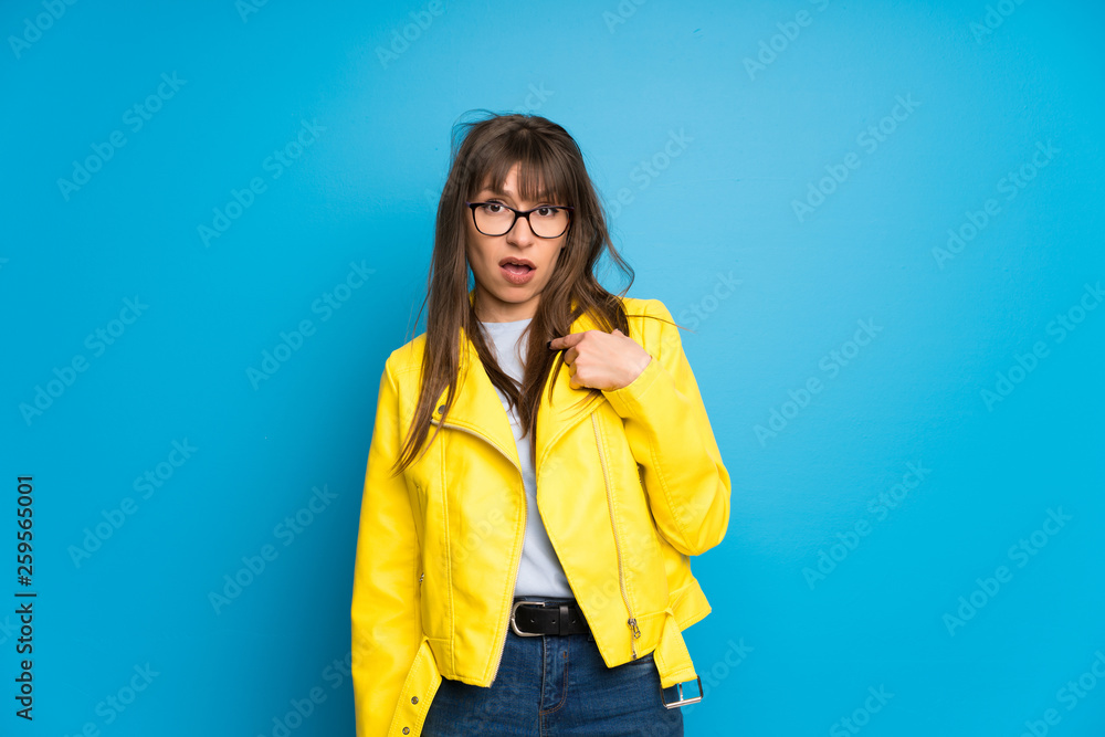 Young woman with yellow jacket on blue background surprised and shocked while looking right Young woman with yellow jacket on blue background surprised and shocked while looking right