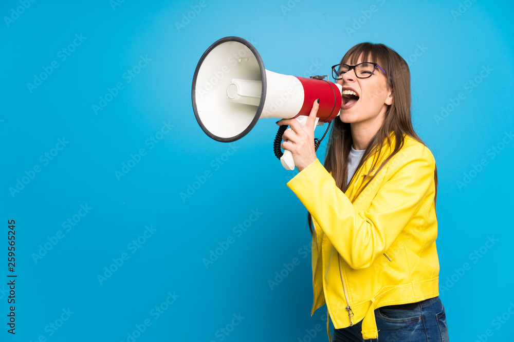 Young woman with yellow jacket on blue background shouting through a megaphone Young woman with yellow jacket on blue background shouting through a megaphone