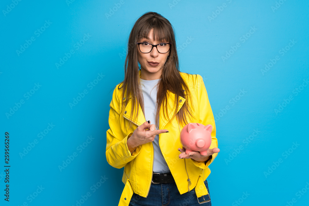 Young woman with yellow jacket on blue background holding a piggybank Young woman with yellow jacket on blue background holding a piggybank