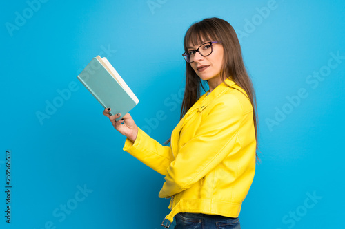Young woman with yellow jacket on blue background holding a book and enjoying reading