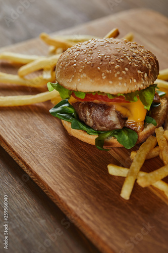 delicious meat burger with cheese, greenery and sesame near french fries on wooden chopping board