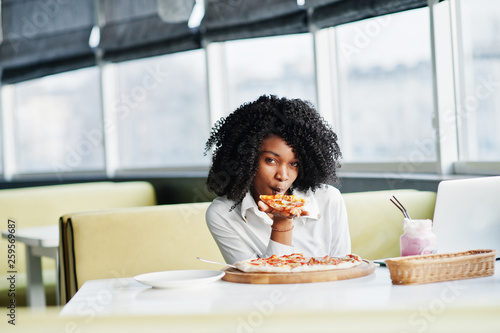 Сheerful business african american lady with afro hair, wear white blouse sitting at table, work with laptop in cafe, eat pizza and drink pink milkshake cocktail.