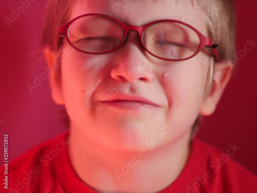 Upset child in red sweater on red background is crying and actively expresses emotions. Close up little kid boy in glasses crying with tearful on her face
