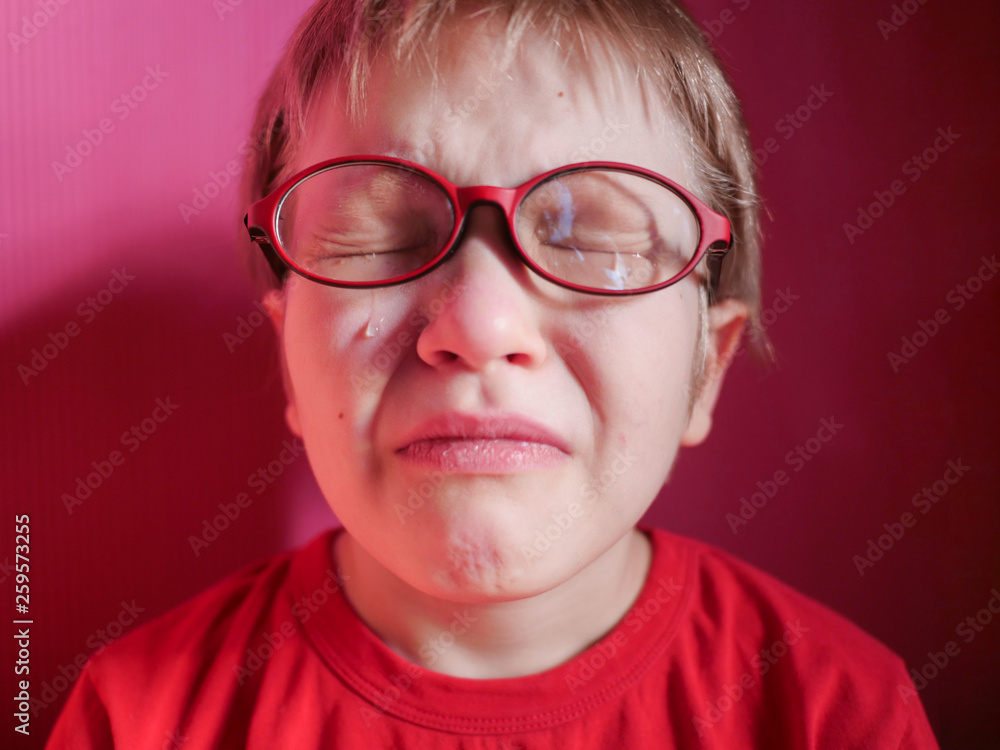 Upset child in red sweater on red background is crying and actively expresses emotions. Close up little kid boy in glasses crying with tearful on her face