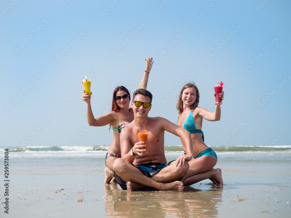 Two girls and a man on the beach drinking juice from glass glasses Two girls and a man on the beach drinking juice from glass glasses