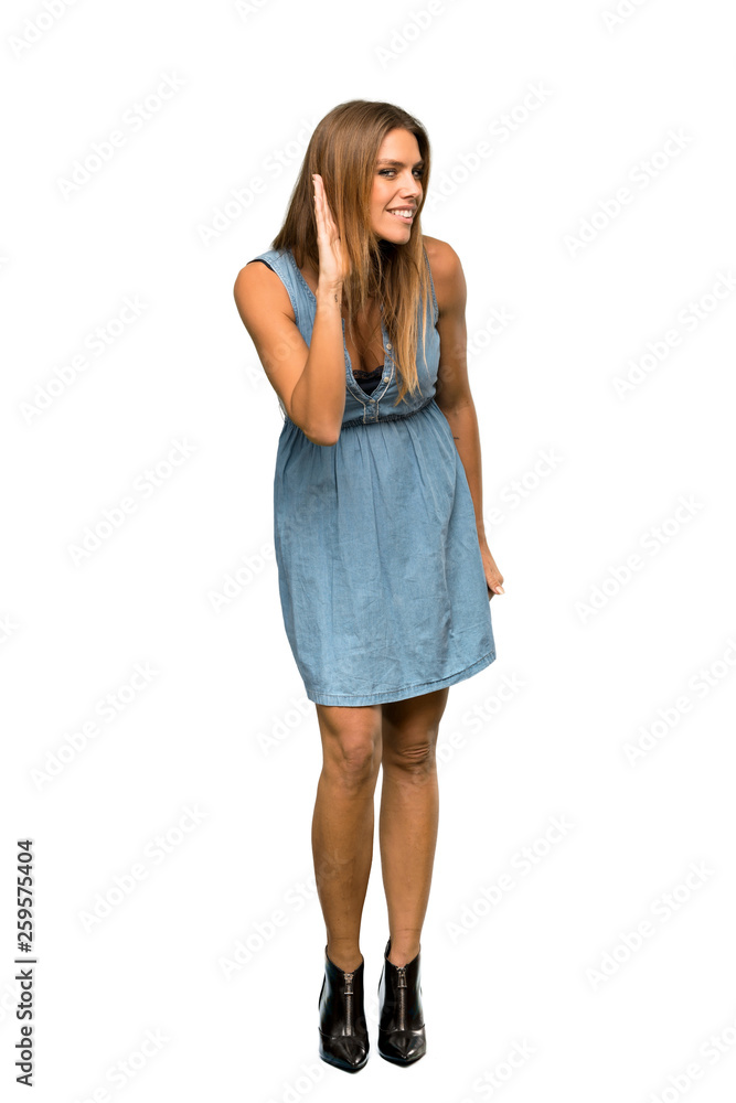 A full-length shot of a Blonde woman with jean dress listening to something by putting hand on the ear over isolated white background A full-length shot of a Blonde woman with jean dress listening to something by putting hand on the ear over isolated white background
