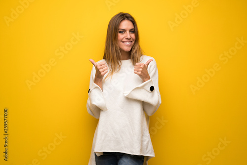 Blonde woman over yellow wall giving a thumbs up gesture with both hands and smiling