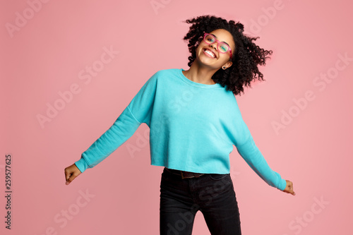 Excited african american young woman with bright smile dressed in casual clothes, glasses and headphones dance over pink background.