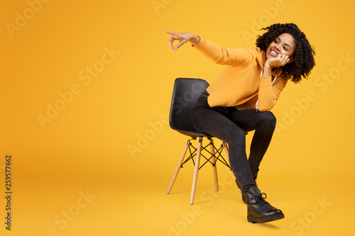 Charming african american young woman with beautiful smile dressed in casual clothes sitting on the stylish chair and point finger away over yellow background.