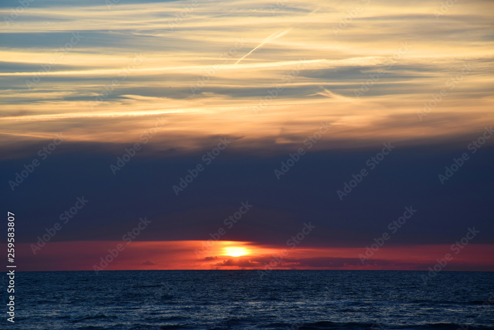 Obraz premium Sunset over the Ocean - View of the red sun sinking into the horizon and waves washing over the sand of the beach