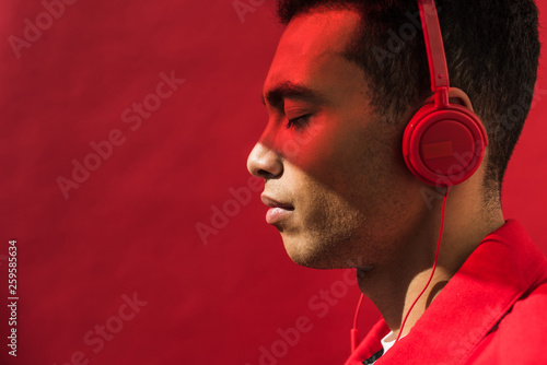 side view of handsome mixed race man in headphones with eyes closed isolated on red with copy space
