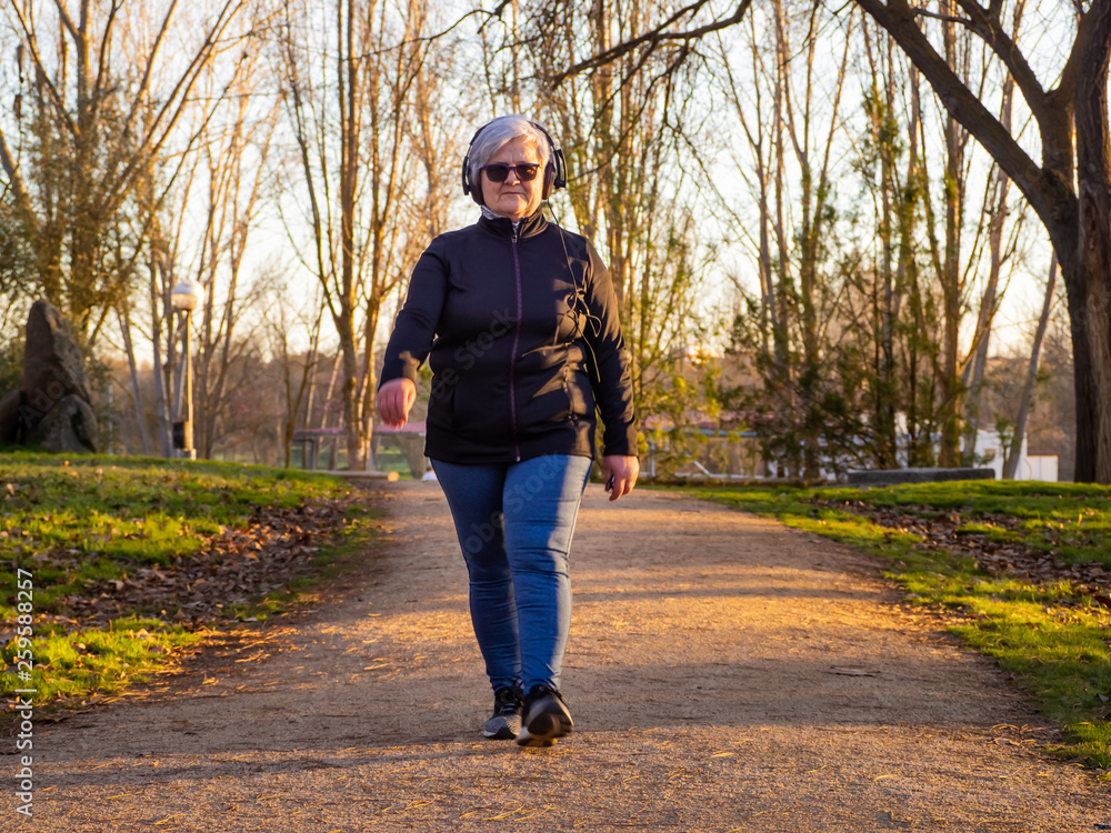 Fototapeta premium A senior woman with white hair listening to music with headphones walking in an urban park