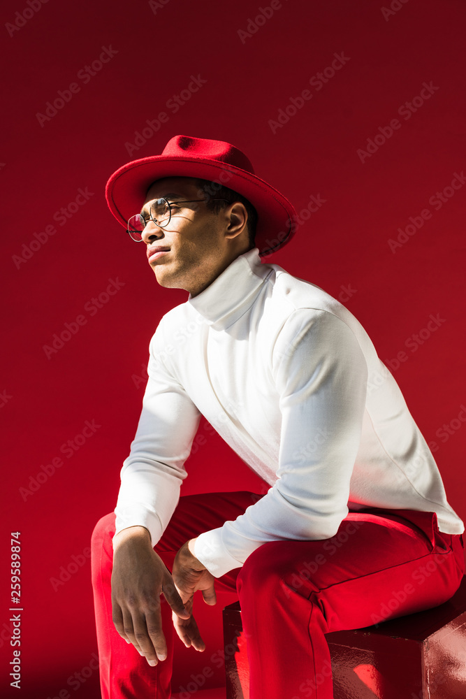 fashionable mixed race man in hat and glasses sitting and posing isolated on red fashionable mixed race man in hat and glasses sitting and posing isolated on red