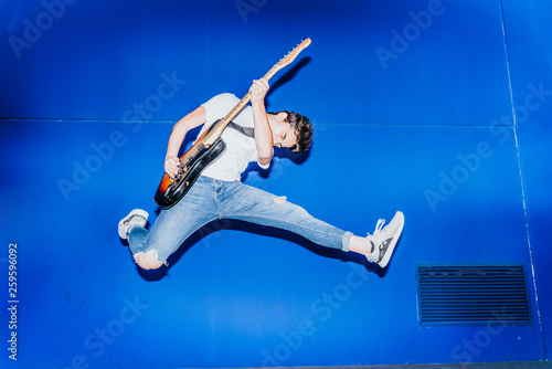 Quadro su tela young man jumping with electric guitar on blue background