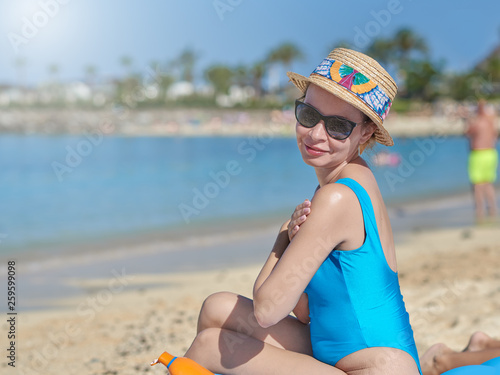 Young lady in a sunhat and sunglasses is covering herself with sun protection lotion, while being at the beach. © Artem