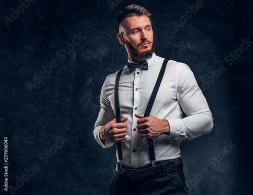 Stylishly dressed young man in shirt with bow tie and suspenders. Studio photo against dark wall background © Fxquadro