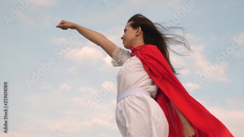 girl dreams of becoming a superhero. beautiful superhero girl standing on a field in a red cloak, cloak fluttering in the wind. Slow motion. young woman plays in red cloak with expression of dreams.