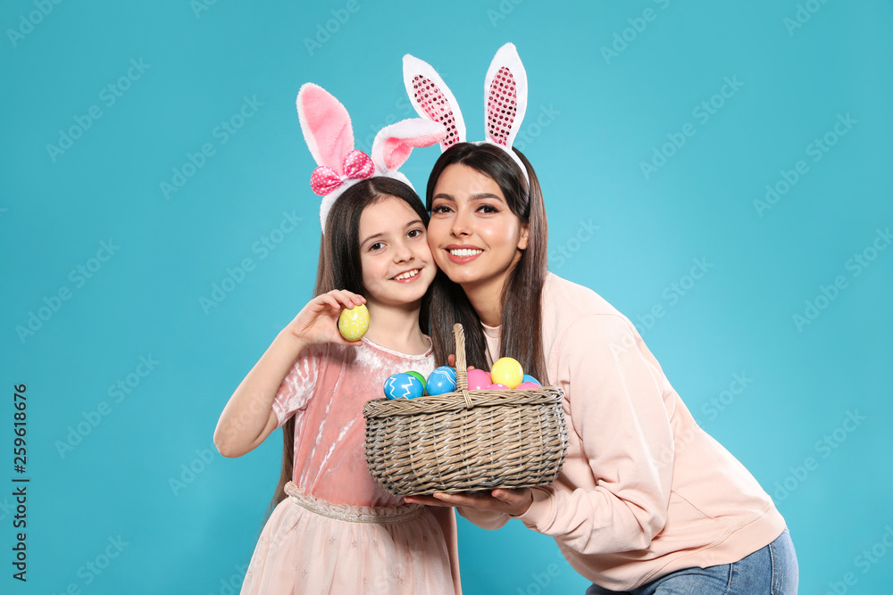 Mother and daughter in bunny ears headbands with basket of Easter eggs on color background Mother and daughter in bunny ears headbands with basket of Easter eggs on color background
