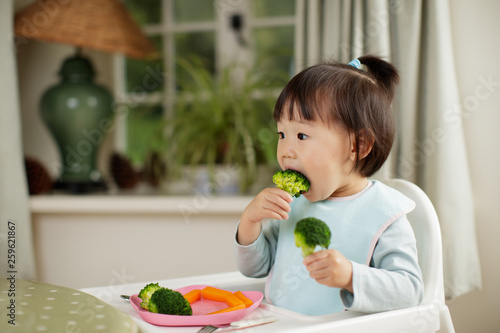 toddler girl eating  healthy  vegetable sitting on high chair beside a dinner table at home