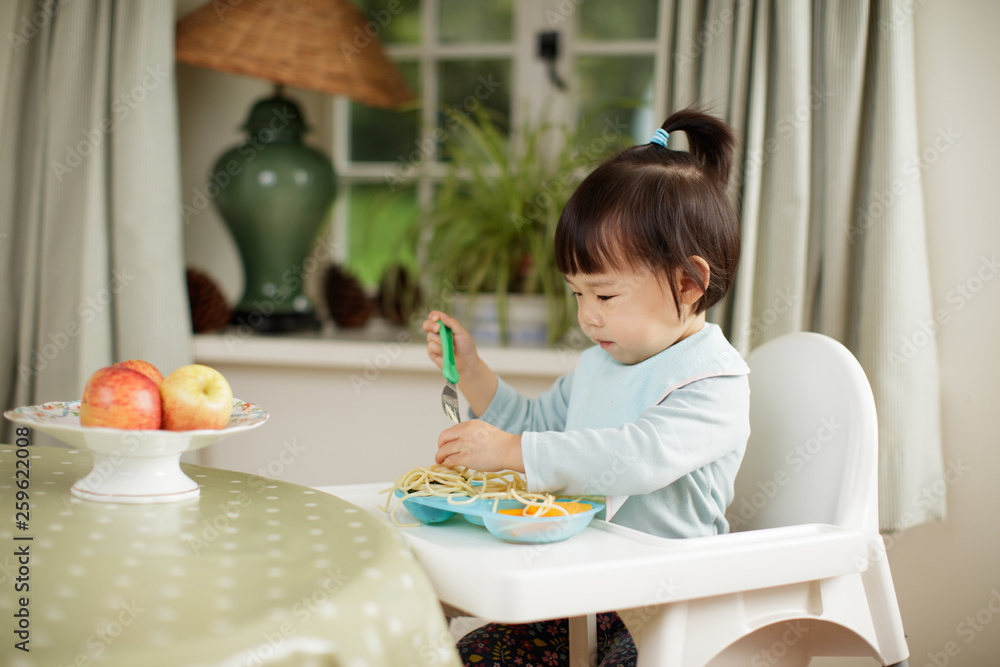 toddler girl eating healthy pasta and vegetable sitting on high chair beside a dinner table at home toddler girl eating healthy pasta and vegetable sitting on high chair beside a dinner table at home