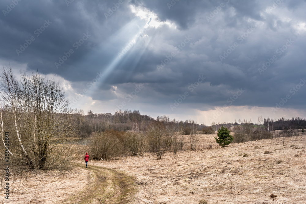a ray of light from the clouds, a woman in a red jacket is walking along a rural road, early spring nature