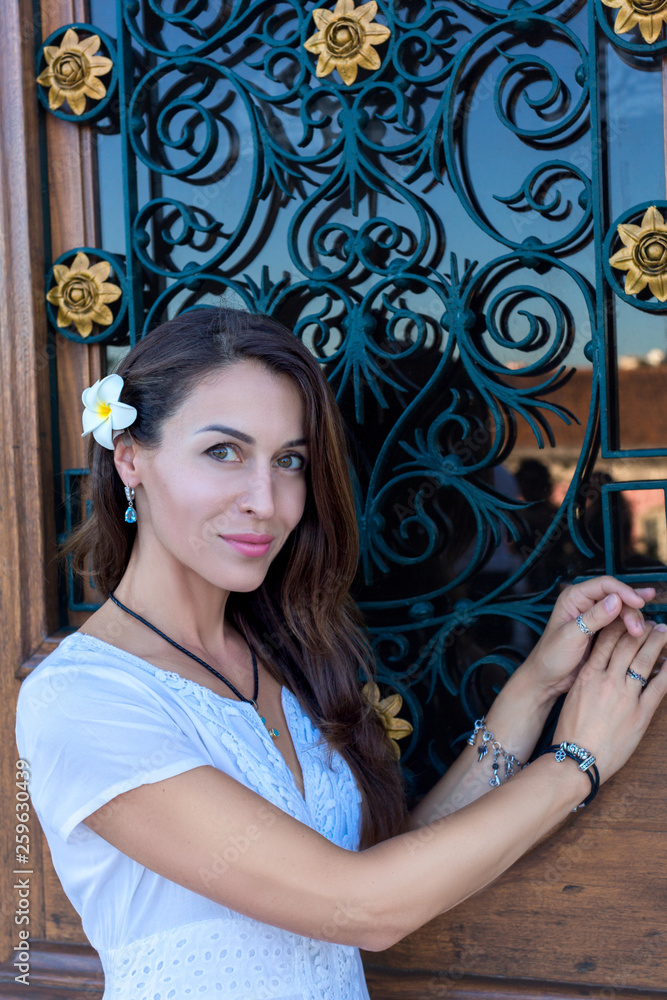 Fototapeta premium Portrait of beautiful girl with long hair in a white dress and a frangipani plumeria flower on the background of a wooden door with glass and wrought-iron bars