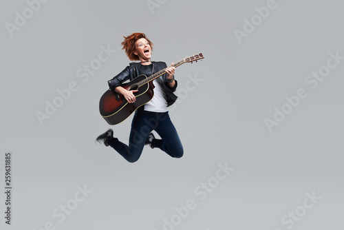 Young woman musician with an acoustic guitar in hand on a gray background. She laughs and jumps high. plays rock and roll loudly. Full-length portrait.