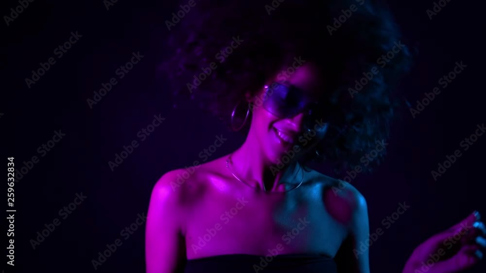 Young african girl with curly hair dancing in neon light. Tempting woman with perfect make-up smiling, enjoying moment, music. Glamour, fashion, style concept Young african girl with curly hair dancing in neon light. Tempting woman with perfect make-up smiling, enjoying moment, music. Glamour, fashion, style concept