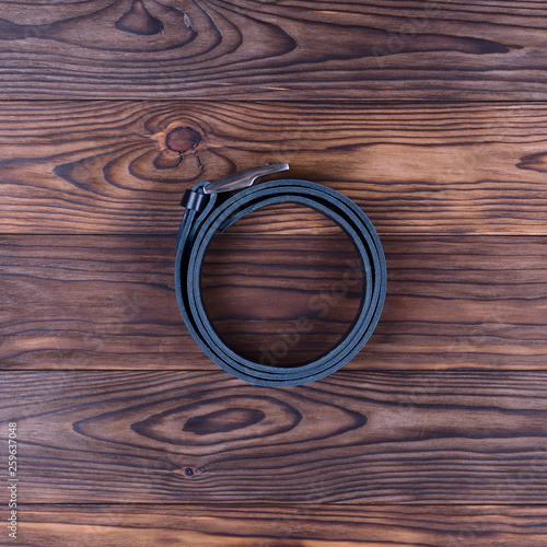 Black color handmade belt lies on textured wooden background. The belt is twisted into a ring. Up to down view. Stock photo of businessman accessories.