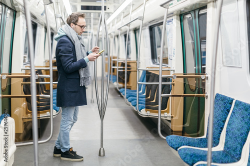 Male with glasses and wireless headphones rides in the subway train. He is wearing a blue coat and a gray scarf, holding handrail and using smartphone. Digital dependence, social networks addiction.