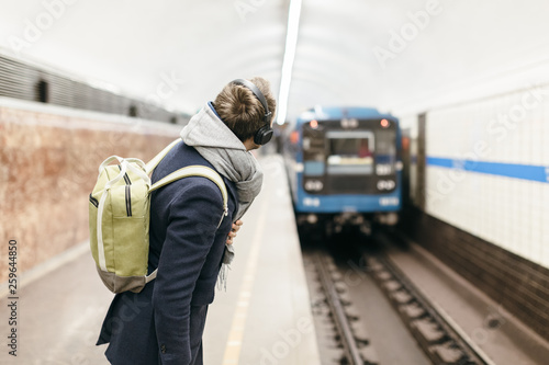 A young handsome man in a blue coat with green backpack behind and headphones on the head looks after the departing metro train. Selective focus on the male. Copy space. Concept of time.